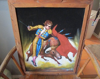May include: A velvet painting in a wooden frame depicting a matador in a gold and blue suit of lights holding a red cape while facing a brown bull. The matador holds a sword in one hand, and the scene is set against a dark black background.