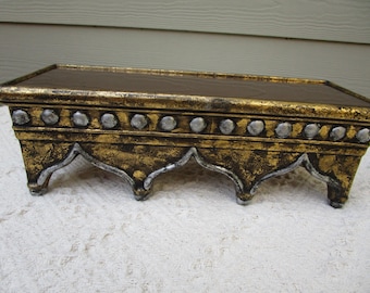 May include: A rectangular, decorative shelf with a dark brown top and ornate gold and silver painted trim. The shelf has a distressed, antique appearance, with decorative silver accents along the front edge and curved legs.