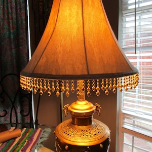 May include: Ornate brass table lamp with a textured, amber-colored lampshade. The lamp features intricate detailing on the base and a beaded fringe. The lamp is a decorative lighting fixture.