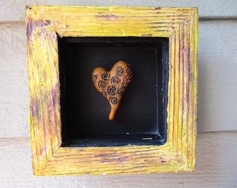 May include: A square, textured yellow frame with a black interior shadow box. Centered inside is a small, tan-coloured heart ornament featuring a dark, hand-drawn spiral pattern across its surface. The frame has a distressed, layered paint finish.