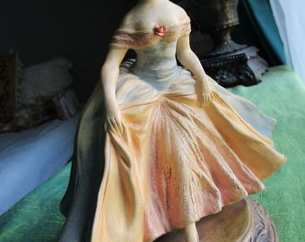 May include: A ceramic figurine of a person wearing an off-the-shoulder gown with a small rose accent at the neckline. The dress features a gradient of peach, yellow, and blue tones with a textured skirt. The figure stands on a round, ornate base.
