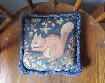 May include: A square decorative pillow featuring a woven tapestry design of a light pink squirrel sitting among green foliage and small flowers on a dark background. The pillow is finished with a textured blue fringe trim along all four edges.