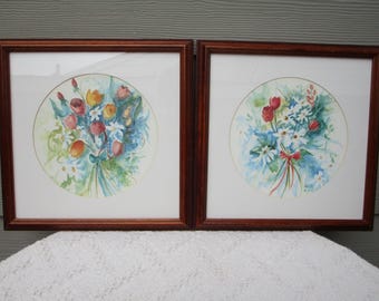 May include: Two framed watercolor floral prints. Each print features a bouquet of flowers in a circular design. The flowers are in shades of red, orange, yellow, and blue, with green foliage. The frames are a dark brown color.