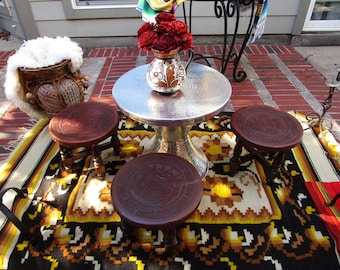 May include: An outdoor setting with a silver metal table and three small, dark wooden stools with carved tops. A vase of red roses sits on the table. The scene is set on a patterned rug. The table is approximately 76 cm in diameter.