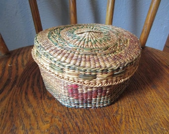 May include: A round, lidded basket crafted from woven natural fibres in shades of tan, green, and brown. The exterior features a subtle, integrated pattern of red and dark green accents woven into the circular lid and the sides of the container.