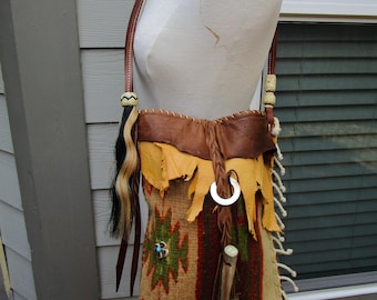 May include: A handcrafted shoulder bag with a Native American design. The bag features a woven fabric with a geometric pattern in shades of brown, green, and red. It has a leather strap, fringe, and a decorative silver ring. The bag is displayed on a mannequin.