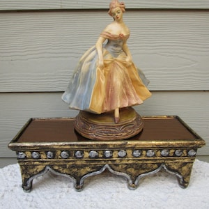 May include: A porcelain figurine of a woman in a flowing gown, in shades of blue, yellow, and peach, stands on a decorative base. The figurine is displayed on a rectangular, ornate stand with gold and silver accents, adding a touch of elegance.