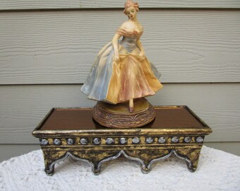 May include: A porcelain figurine of a woman in a flowing gown, in shades of blue, yellow, and peach, stands on a decorative base. The figurine is displayed on a rectangular, ornate stand with gold and silver accents, adding a touch of elegance.
