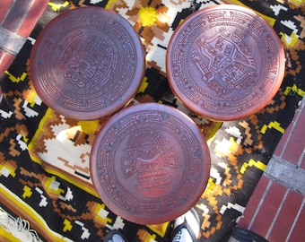 May include: Three round, reddish-brown ceramic plates with intricate, embossed designs. Each plate features a different ancient Aztec or Mayan-inspired motif. The plates are arranged on a colorful, patterned textile.