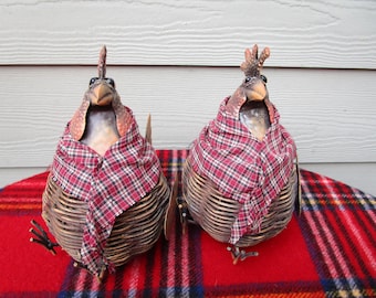 May include: Two decorative chicken figurines. Each chicken is made of woven wicker and wears a red and black plaid scarf. The chickens have detailed beaks, eyes, and crests. They are sitting on a red plaid blanket.