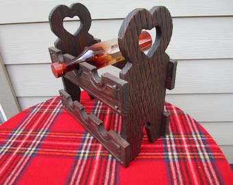 May include: A dark brown wooden wine rack with heart-shaped cutouts. The rack holds a brown glass bottle with a red stopper. The rack is on a red plaid blanket.