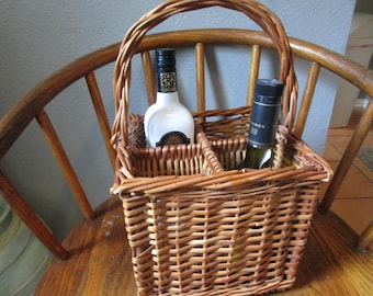 May include: A rectangular brown woven wicker basket with a tall arched handle and two internal compartments. One compartment holds a white bottle with a black cap and label, while the other contains a dark glass bottle with a black screw-top cap.