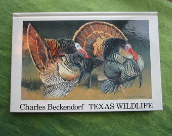 May include: A hardback book titled Charles Beckendorf TEXAS WILDLIFE featuring a detailed painting of two wild turkeys with fanned tail feathers, dark plumage, and red wattles on a white background with black text at the bottom of the cover.