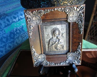May include: A framed religious icon featuring a silver relief of the Virgin Mary and child. The icon is set within a dark wood frame with ornate silver corner embellishments. The frame is supported by a small black stand.