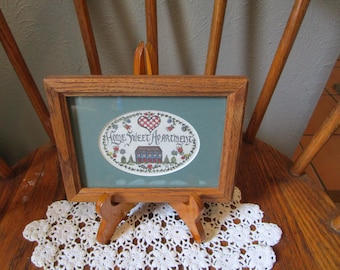 May include: A rectangular wooden frame containing an oval-shaped print with a sage green mat. The print features the text "Home Sweet Apartment" in black script, a red and white checkered heart, and a small illustration of a red house with trees.