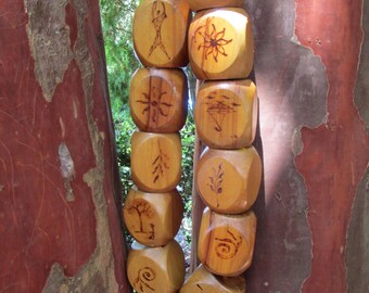 May include: A vertical strand of light brown wooden beads, each featuring a unique dark brown pyrography design. The symbols include a sun, a fish, a flower, a tree, a spiral, and various abstract patterns, all strung together on a thin cord.