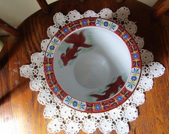May include: A white ceramic bowl featuring two painted red goldfish swimming inside. The rim is decorated with a wide band of red, blue, and gold floral patterns. The interior of the bowl is smooth and white, providing a clear view of the fish.