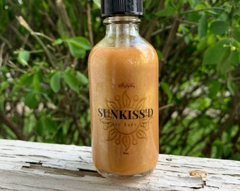 May include: A glass bottle with a gold liquid and a black dropper top. The bottle has a gold label with the text "SUNKISS'D Summer Body", a sun graphic, and "2 oz".