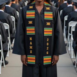 May include: A black graduation gown with a decorative sash. The sash is black with horizontal stripes of green, yellow, and red. The word "NAME" is vertically printed on one side, and the year "2026" is on the other.