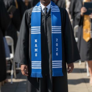 以下が含まれることがあります： 白のストライプと片面に縦書きで「NAME」、もう片面に「2026」とプリントされた青い卒業ストール。黒い卒業ガウンの上から着用。ストールにはフリンジエッジが付いています。