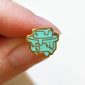 May include: A teal enamel pin with a gold outline. The pin features a stylized cake design with a top layer and dripping frosting. The pin is small and likely intended for clothing or accessories.