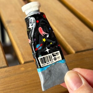 Puede incluir: Un tubo de suministro de arte en miniatura, pintado a mano, con un dise&ntilde;o c&oacute;smico. El tubo presenta un fondo negro con planetas, estrellas y nebulosas coloridas. La tapa es blanca y la base es gris. El tubo es sostenido por una persona.