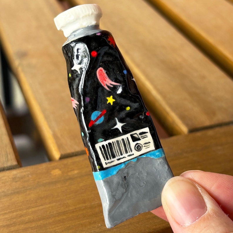Puede incluir: Un tubo de suministro de arte en miniatura, pintado a mano, con un dise&ntilde;o c&oacute;smico. El tubo presenta un fondo negro con planetas, estrellas y nebulosas coloridas. La tapa es blanca y la base es gris. El tubo es sostenido por una persona.