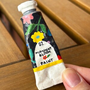 Puede incluir: Un peque&ntilde;o tubo de pintura pintado a mano, con una tapa blanca y un cuerpo azul oscuro. El tubo est&aacute; decorado con flores coloridas y las palabras "Midnight Bloom Paint". El n&uacute;mero "92" tambi&eacute;n es visible.