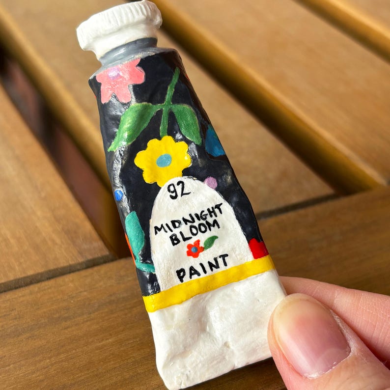 Puede incluir: Un peque&ntilde;o tubo de pintura pintado a mano, con una tapa blanca y un cuerpo azul oscuro. El tubo est&aacute; decorado con flores coloridas y las palabras "Midnight Bloom Paint". El n&uacute;mero "92" tambi&eacute;n es visible.