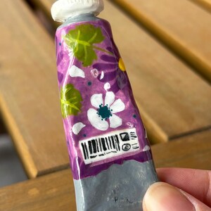 Puede incluir: Un tubo de pintura morado en miniatura, pintado a mano, con dise&ntilde;os florales blancos y hojas verdes. El tubo tiene una base gris y una tapa blanca. Un c&oacute;digo de barras y otras marcas son visibles.