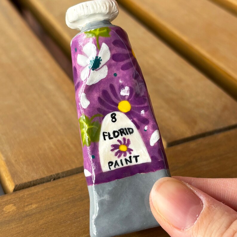 Puede incluir: Un tubo de pintura de cer&aacute;mica con un dise&ntilde;o floral p&uacute;rpura y el texto "8 FLORID PAINT". El tubo tiene una tapa blanca y una base gris. El dise&ntilde;o incluye flores blancas y hojas verdes.