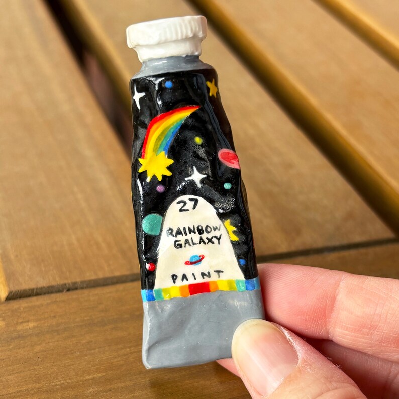 Puede incluir: Un tubo de pintura de cer&aacute;mica con fondo negro, un arco&iacute;ris y la inscripci&oacute;n "27 Rainbow Galaxy Paint". El tubo tiene una tapa blanca y una base gris. El dise&ntilde;o incluye estrellas y planetas.