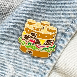 Puede incluir: Un colorido pin esmaltado con forma de hamburguesa apilada. El pin presenta capas de pan, hamburguesa, queso, lechuga, tomate y cebolla. El pin tiene un contorno dorado y está sobre un fondo de mezclilla.