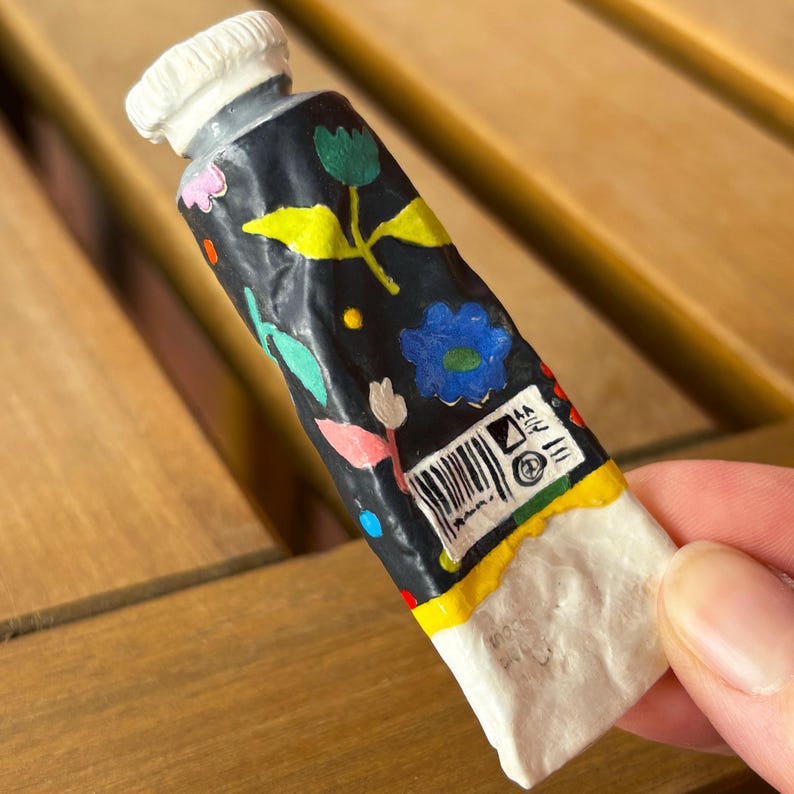 Puede incluir: Un tubo de pintura negro decorado con dise&ntilde;os florales coloridos, que incluyen flores azules, rosas y amarillas, y hojas verdes. El tubo tiene una tapa blanca y una base blanca. El tubo es sostenido por una persona.