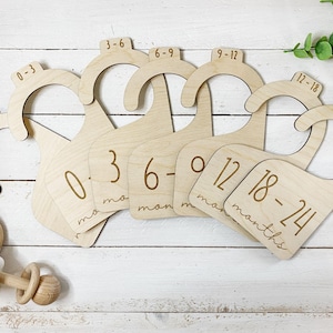 Puede incluir: Conjunto de perchas de madera grabadas con números y texto que indican rangos de edad para bebés, desde recién nacidos hasta 24 meses. Las perchas están hechas de madera natural y tienen un diseño simple y minimalista.