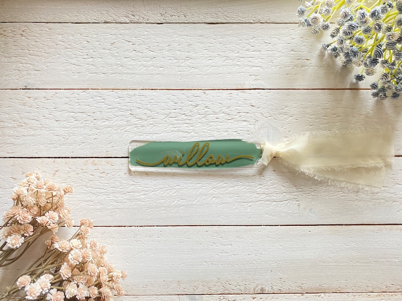 Peut inclure: Un marque-page en verre vert avec le mot "willow" &eacute;crit en lettres dor&eacute;es. Le marque-page est attach&eacute; &agrave; un ruban blanc.