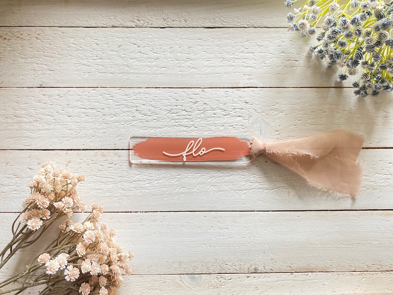 Peut inclure: Un marque-page en acrylique transparent avec une bande peinte de couleur p&ecirc;che et le mot "flo" &eacute;crit en blanc. Un ruban rose clair est attach&eacute; au marque-page.