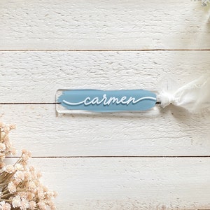 Peut inclure: Une carte de nom en acrylique bleu clair avec le nom "carmen" &eacute;crit en cursive blanche. La carte est attach&eacute;e avec un ruban blanc.