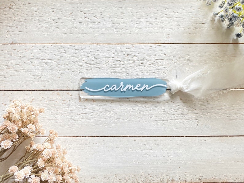 Peut inclure: Une carte de nom en acrylique bleu clair avec le nom "carmen" &eacute;crit en cursive blanche. La carte est attach&eacute;e avec un ruban blanc.