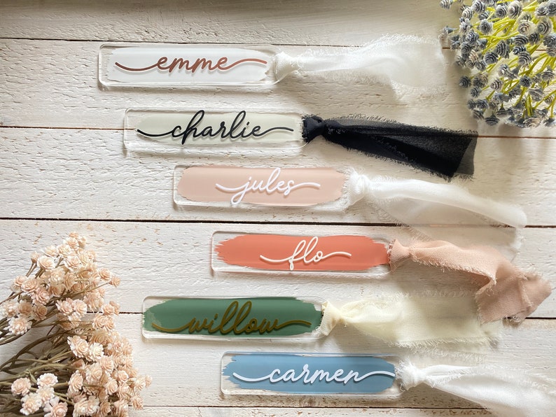 Peut inclure: Six &eacute;tiquettes de nom en acrylique avec des fonds de couleurs diff&eacute;rentes et des lettres blanches ou dor&eacute;es. Les noms sont "Emme", "Charlie", "Jules", "Flo", "Willow" et "Carmen". Chaque &eacute;tiquette est accompagn&eacute;e d'un ruban assorti.