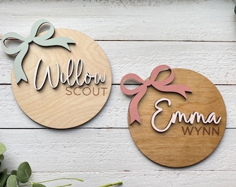 May include: Two wooden name plaques. One has a light green bow and the name "Willow SCOUT" in white and brown. The other has a pink bow and the name "Emma WYNN" in white and brown. Both are circular.