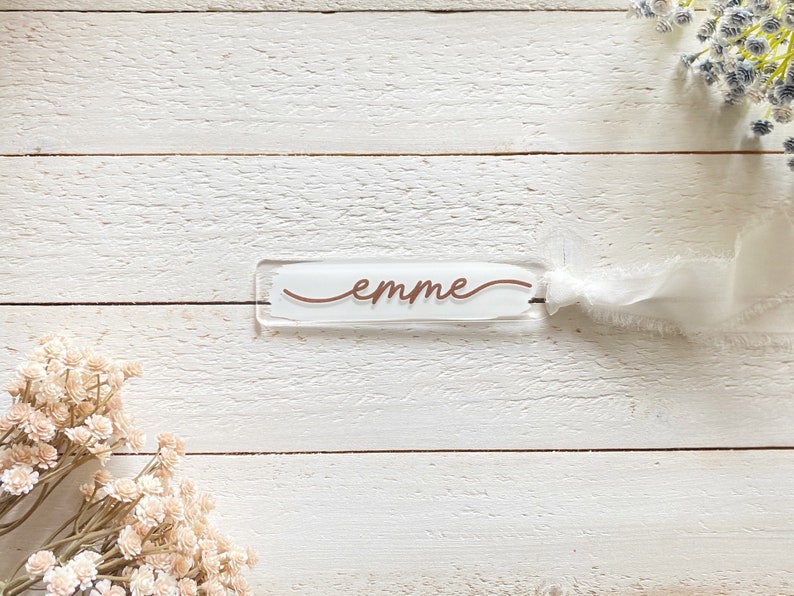 Peut inclure: Un marque-page en acrylique blanc avec le mot "emme" &eacute;crit en cursive en cuivre.