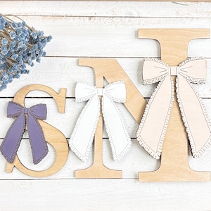 May include: Wooden letters spelling "SNM" with decorative bows. The letter "S" has a purple bow, the "N" has a white bow, and the "M" has a light pink bow. The bows have a scalloped edge. The letters are on a white wooden surface.