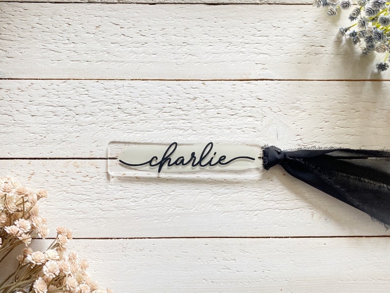 Peut inclure: Un marque-page en acrylique transparent avec le nom "charlie" &eacute;crit en cursive noire. Le marque-page est attach&eacute; &agrave; un ruban noir.
