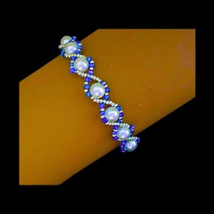 May include: A beaded bracelet with white faux pearls and blue accents. The bracelet is displayed on a tan surface against a black background. The design features a repeating pattern of pearls and blue beads.
