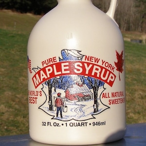 May include: A cream-colored jug of New York maple syrup with a black cap and handle. The label features a red maple leaf and the words "Pure Maple Syrup" in a red banner. The jug holds 32 fl oz, or 1 quart, of syrup.