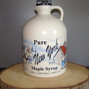 May include: A white ceramic jug with a black lid and a handle. The jug is decorated with a winter scene featuring a horse-drawn sleigh, a farmhouse, and the words "Pure New York Maple Syrup". The jug is labeled with the size "1.89 L - Half Gallon - 64 Fl. Oz."
