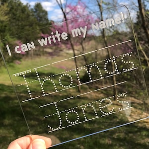 May include: A clear plastic dry-erase board with the text "I can write my name!" at the top. The board has dotted lines for tracing the names "Thomas" and "Jones".