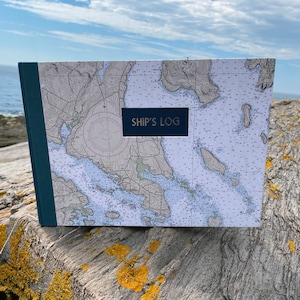 May include: A blue and white nautical chart themed journal with the text "SHIP'S LOG" on the cover. The journal is sitting on a weathered piece of driftwood.