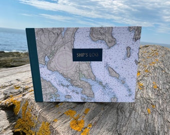 May include: A blue and white nautical chart themed journal with the text "SHIP'S LOG" on the cover. The journal is sitting on a weathered piece of driftwood.